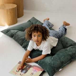 Child lying on a green tufted corduroy cushion reading a book in a cozy room.