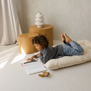 Child lying on a French style cushion reading a book in a cozy room with wooden stools and a vase.