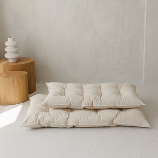 Two Beige tufted floor cushions with wooden side tables and a vase in the background