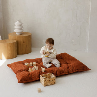 Child playing on a large tufted corduroy floor cushion with wooden blocks and a wicker basket in a minimalistic room.