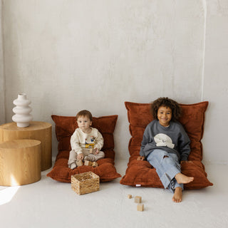 Two children sitting on large terracotta floor cushions in a minimalistic room with wooden tables and toys.