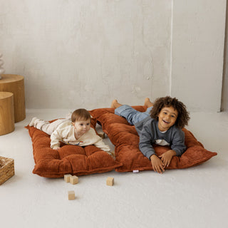 Two children playing on a large terracotta tufted floor cushion with wooden blocks around them.