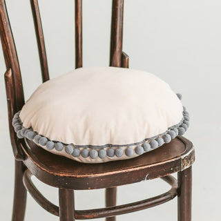 Round beige cushion with grey pompom trim styled on a chair